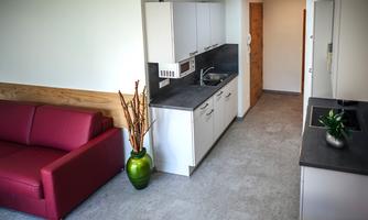 A modern kitchen with white cabinets and a gray countertop. In the foreground, there is a red sofa and a green plant.