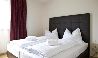 A cozy bedroom with a large double bed and elegant bedding. The walls are light, and curtains in dark red complement the decor.