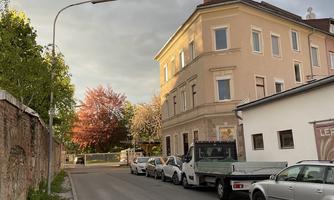 A quiet street with parked cars and a historic building. In the background, green trees and some colorful leaves are visible.