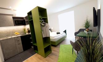 A modern apartment kitchen with an open living area. The decor is minimalist and features a green shelving wall as well as a sleeping area.