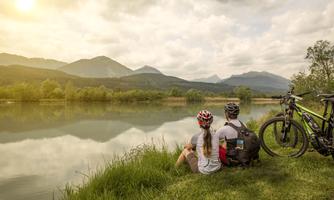 A couple sits by the shore of a lake and looks at the mountains. Two bicycles are standing next to them in the grass.
