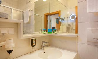 A modern bathroom with a sink and a large mirror. The walls are light tiled and there are towels as well as toiletries.