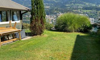 A green meadow with a view of mountains and a city in the background. A cozy house with terrace furniture is also visible.