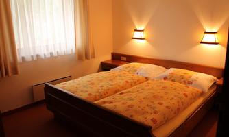 A cozy bedroom with a double bed and colorful bedspreads. The room has windows with curtains and soft lighting.