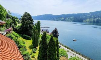 A picturesque landscape with a clear lake and green trees. In the background, gentle hills stretch under a blue sky.