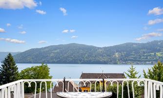 A picturesque view from a balcony of a lake, surrounded by green mountains. The sky is clear and blue, and the landscape is peaceful.