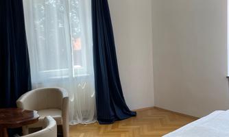 A bright room with a bed, a small table, and two chairs. Large windows with curtains provide plenty of daylight.