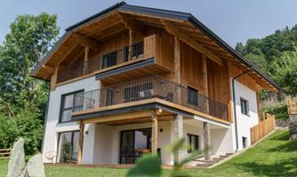 A modern wooden house with a large balcony and a beautiful garden. Surrounded by green trees, it offers a relaxed atmosphere.