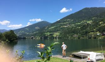 A calm lake surrounded by mountains and lush vegetation. In the foreground, there is a person at the shore looking at a boat.