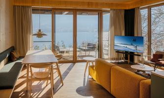 A modern living room with large windows and a view of the water. It is bright and cheerfully furnished with a dining table, a comfortable couch, and a television.