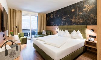 A modern hotel room with a large double bed and a stylish wall design. Large windows provide daylight and access to a balcony.