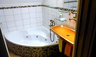 Corner whirlpool tub with massage jets and mosaic cladding, orange sink with gold rim and brass fittings, mirror and glass shelves on a light tiled wall.