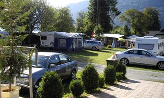 A campsite with several motorhomes and cars in a green setting. In the background, mountains and trees are visible.