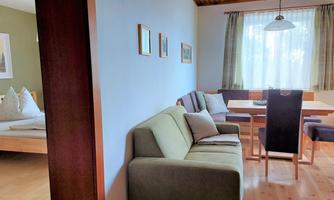 A cozy living space with a sofa and a dining table. The room is bright and cheerful, with large windows and beautiful pictures on the wall.