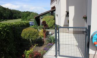A well-tended garden with a low fence and an open driveway. In the background, green trees and a bright house are visible.