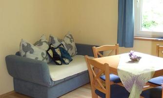 A cozy living area with a blue sofa and a dining table. The room is bright and cheerful, with large windows and a small table.