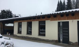A modern house with wooden cladding and large windows. It is situated in a snowy environment with pine trees in the background.