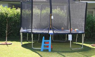 A trampoline with a safety net in the garden. Next to it, there is a small blue staircase and some balls are lying on the trampoline.