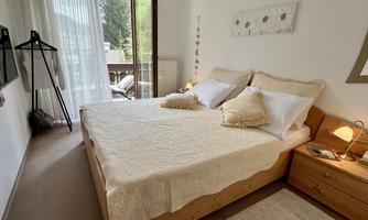 A cozy bedroom with a double bed and white pillows. Large windows offer a view of a balcony and the outdoor area.