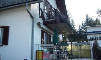 A modern house with a balcony and a seating area. Trees are visible in the background.