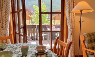A cozy dining area with a table set with dishes overlooks a balcony and the surrounding landscape through large windows. The decor conveys a warm, inviting ambiance.