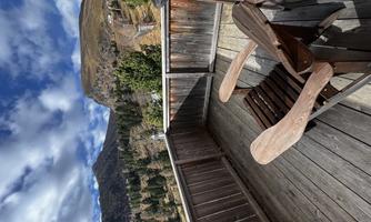 A terrace with a wooden chair and a view of impressive mountains. The sky is partly cloudy and the surrounding area is surrounded by trees.