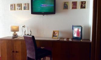 A cozy room with a modern TV, a wooden desk, and a chair. On the wall are picture frames, and a lamp is on the table.