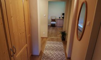 A corridor with wooden cabinets and a round mirror on the wall. At the end of the corridor, a bright room with a chair and a table is visible.