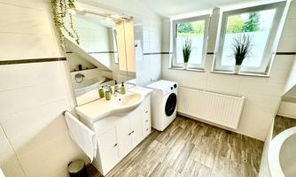 A modern bathroom with a washing machine and a sink. Large windows provide plenty of daylight and plants add a fresh touch.
