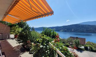 A sunny terrace with colorful awnings and a view of a tranquil lake. The surroundings are green and inviting.