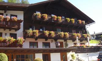 A charming multi-story building with balconies adorned with colorful flowers. The surroundings are enveloped by green mountains, radiating an inviting atmosphere.