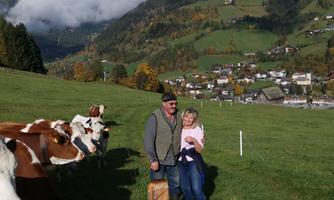 A man and a girl are standing on a meadow with cows in the foreground. In the background, picturesque mountains and a village can be seen.