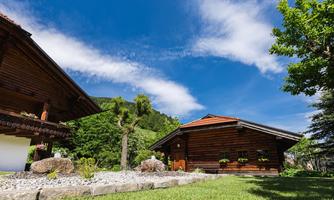 Two traditional wooden houses surrounded by green grass and trees. The sky is clear and blue with some clouds.