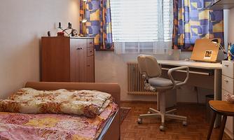A cozy bedroom with a bed and a desk. The windows are adorned with colorful curtains and the room has a warm wooden floor.