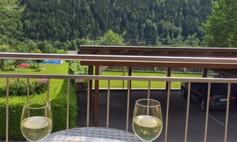 A balcony with a table on which a plate with bread, cheese, and vegetables is placed. Two glasses of wine are next to it, while in the background the mountains and a green landscape can be seen.