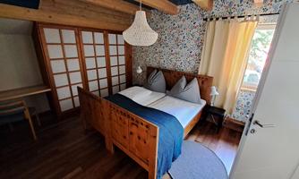 A cozy bedroom with a wooden bed and two pillows. The walls are decorated with a colorful pattern and there is plenty of natural light.