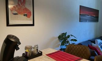 A cozy kitchen with a wooden work surface and a coffee machine. In the background, there are plants and a sofa, as well as a piece of artwork on the wall.