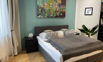 A stylish bedroom with a large bed, decorative cushions, and a cozy throw. A colorful painting hangs on the wall, and a plant stands in the room.
