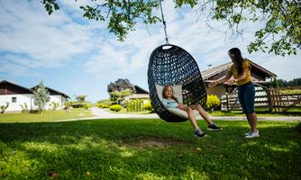 A child is sitting in a swinging wicker chair outdoors. Another person is gently swinging the chair in a beautifully designed garden landscape.