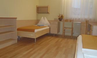 A simple room with two single beds and light wooden floors. There is a desk, a chair, and shelves.