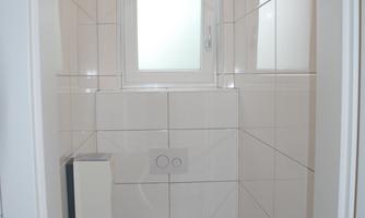 A small bathroom with a toilet and a window. The walls are tiled with white tiles.