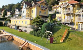 A lovely property with yellow houses and a green garden. In the foreground, a slide and a pier to the water are visible.