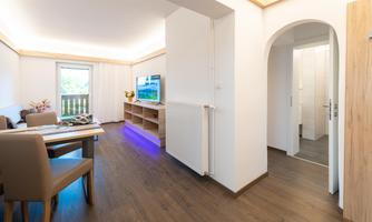A modern living room with a dining area and a large television. The room has wooden flooring and plenty of natural light.