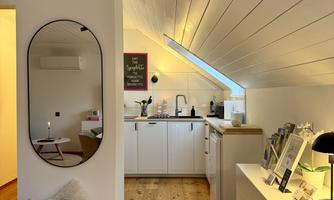 A modern kitchen under the roof with white furniture and wooden floor. A large mirror and a cozy seating area complete the room.
