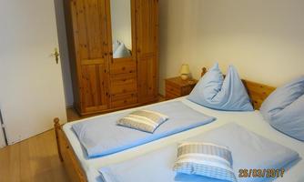 A cozy bedroom with a double bed and two pillows. The wooden wardrobe is reflected in the mirror of the dresser.