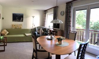 A cozy living area with a dining table, chairs, and a sofa. Large windows let in plenty of natural light and provide a view of greenery.