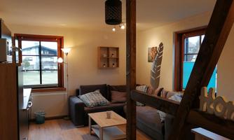 A cozy living room with a sofa, shelves, and a table. Large windows provide plenty of natural light.