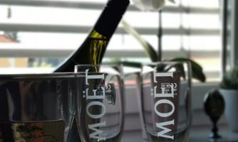 A champagne bottle is standing in a cooler next to two glasses. In the background, an orchid and blinds can be seen.