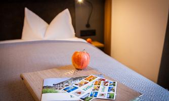 A magazine and some postcards are lying on a bed. A red apple is next to them, while a cozy room is visible in the background.