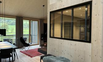 A modern, bright living room with wooden walls and large windows. The room is furnished with a dining table, a chair, and a cozy spot to relax.